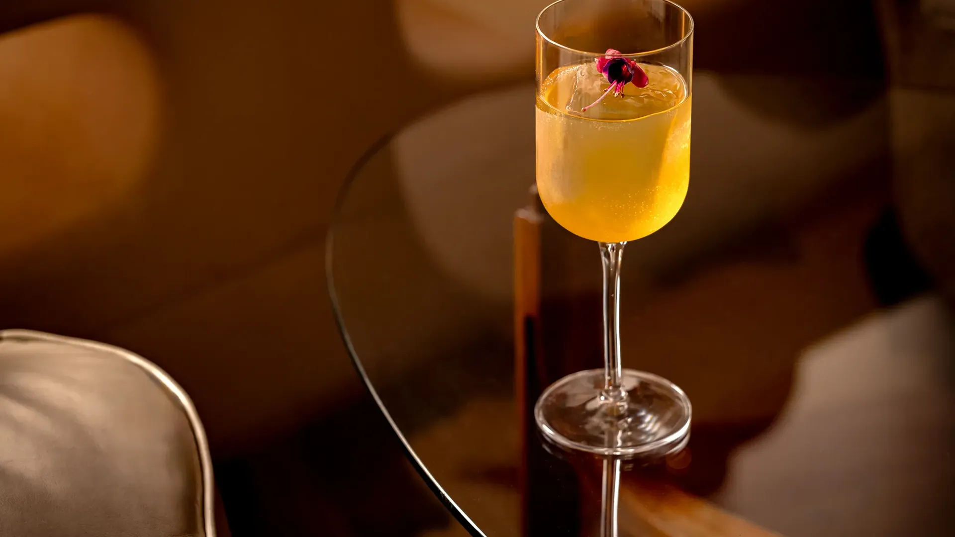 Golden cocktail with flower garnish in stemmed glass on dark wood table.
