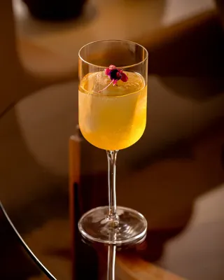 Golden cocktail with flower garnish in stemmed glass on dark wood table.