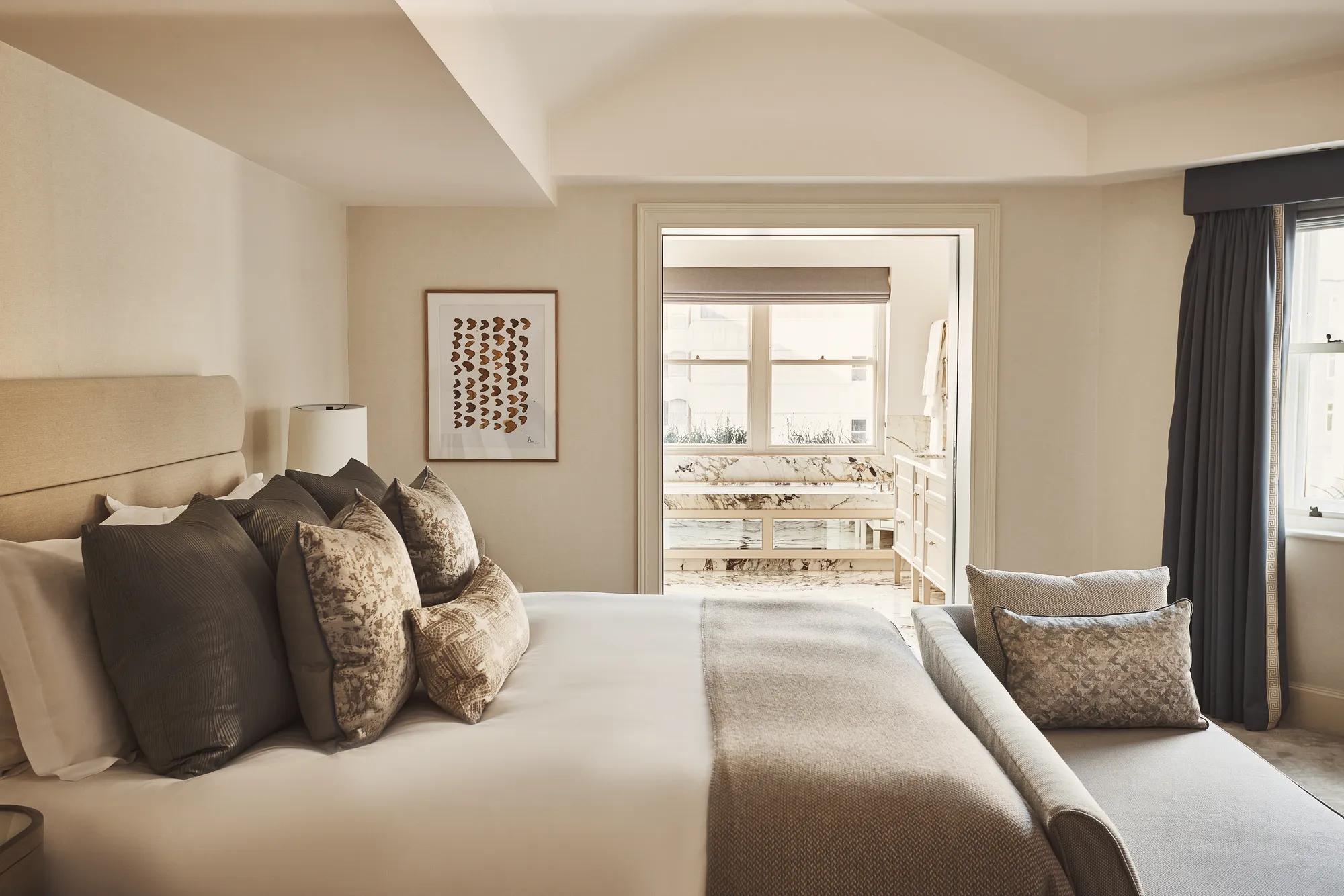 Neutral-toned bedroom with a cushioned bed, bench seating, and a view into a marble bathroom.