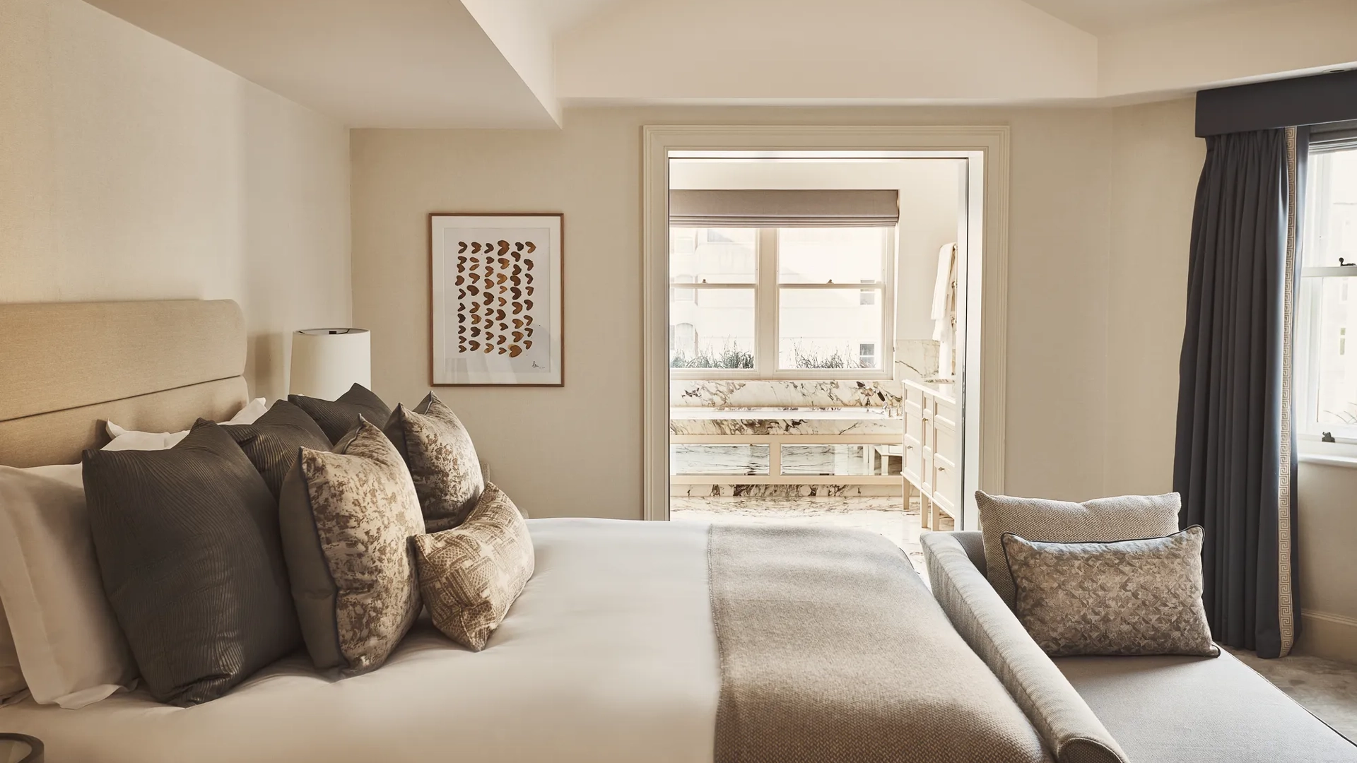 Neutral-toned bedroom with a cushioned bed, bench seating, and a view into a marble bathroom.