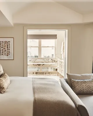 Neutral-toned bedroom with a cushioned bed, bench seating, and a view into a marble bathroom.