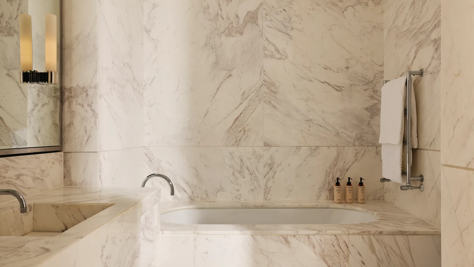 Bathroom clad in white-veined marble, featuring a built-in bathtub, double vanity, and neatly arranged toiletries.