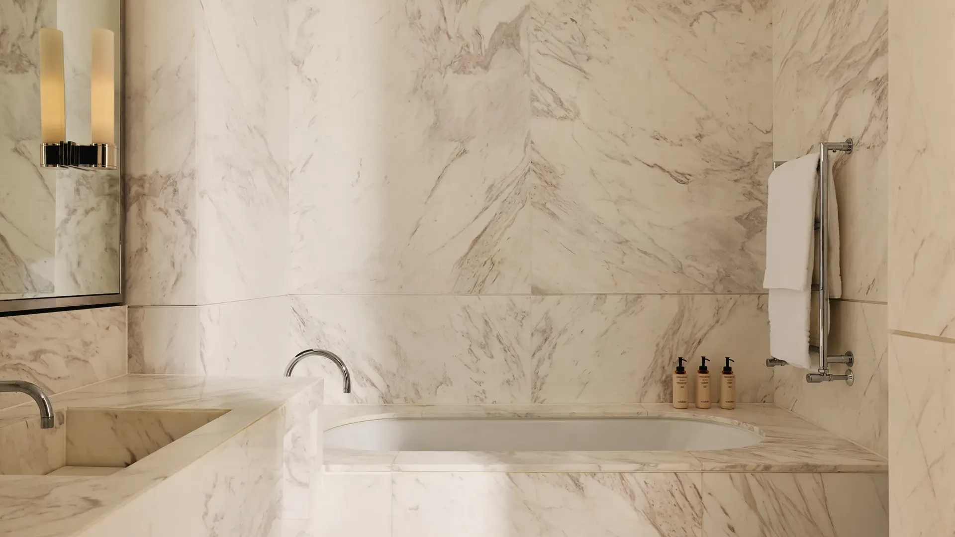 Bathroom clad in white-veined marble, featuring a built-in bathtub, double vanity, and neatly arranged toiletries.