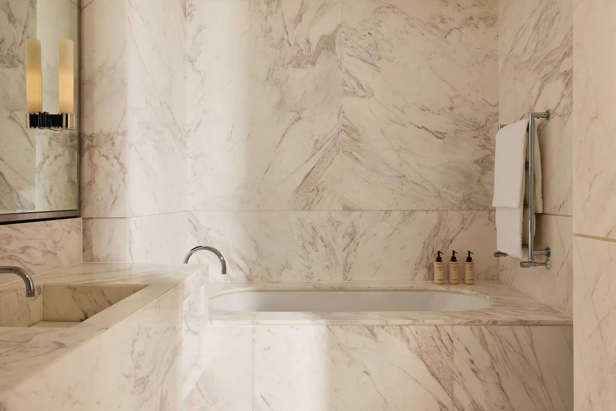 Bathroom clad in white-veined marble, featuring a built-in bathtub, double vanity, and neatly arranged toiletries.