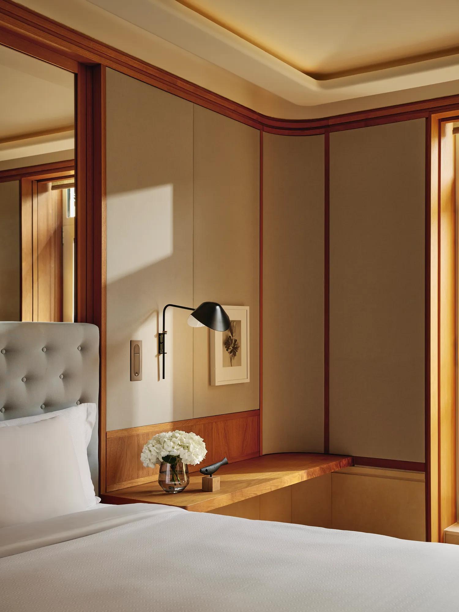 Bedroom corner with a tufted headboard, built-in wooden ledge holding flowers and a phone, and a wall lamp above.