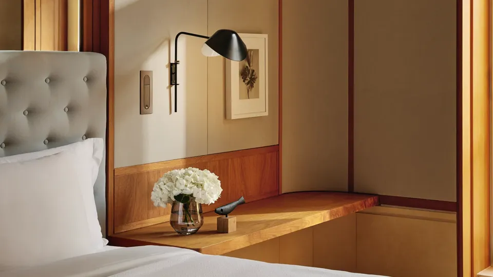 Bedroom corner with a tufted headboard, built-in wooden ledge holding flowers and a phone, and a wall lamp above.