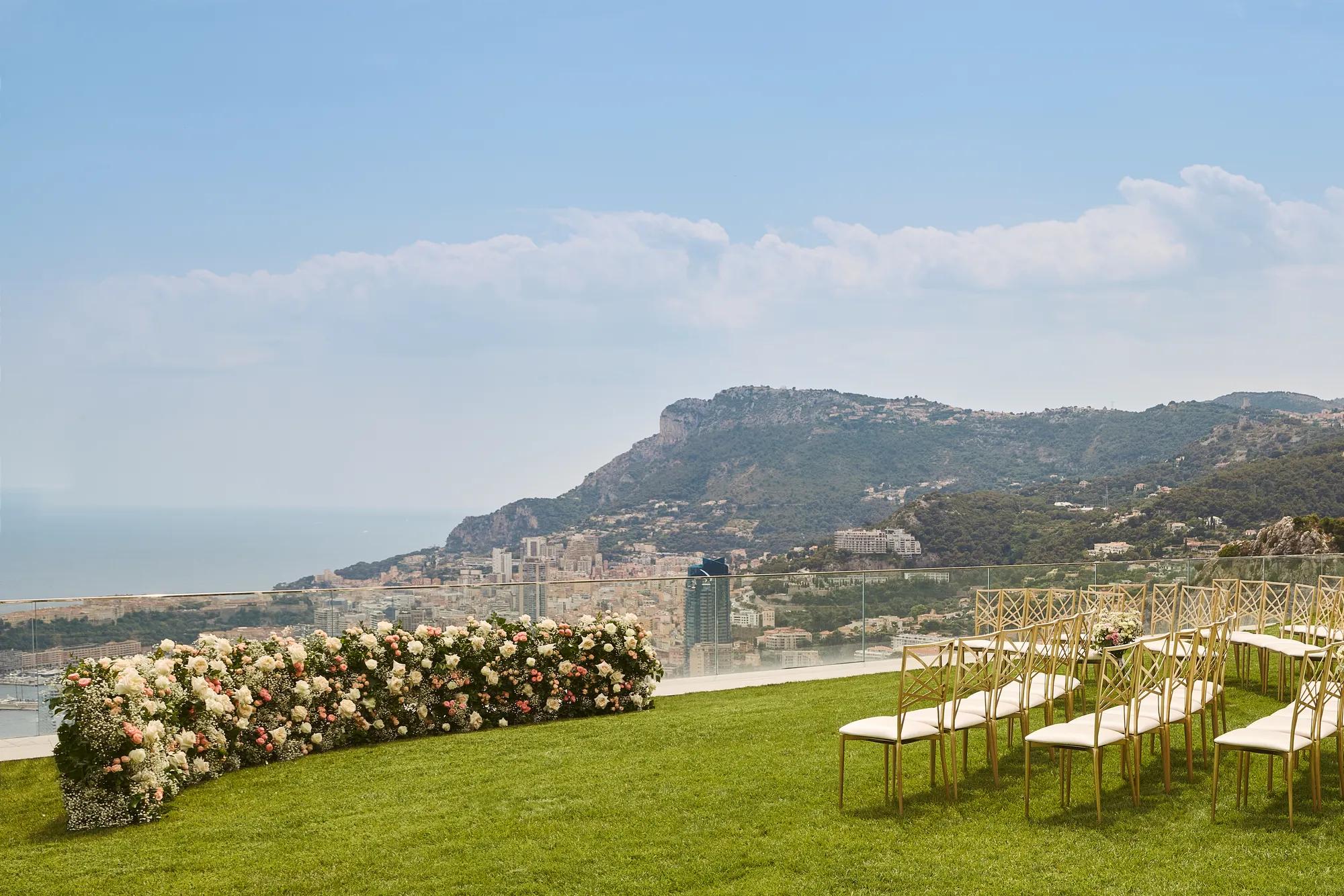 Installation de cérémonie de mariage en plein air avec des rangées de chaises dorées sur une pelouse, face à une composition florale incurvée de roses blanches et roses pâles, avec vue sur la mer et les montagnes.