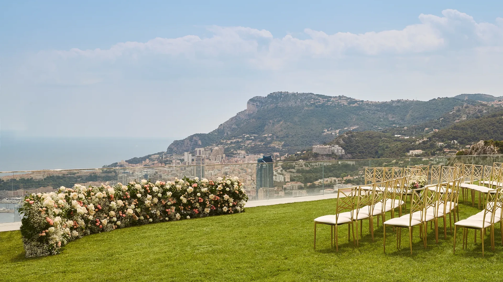 Outdoor wedding ceremony setup with rows of gold chairs on a lawn, facing a curved floral arrangement of white and blush roses, overlooking the sea and mountains.