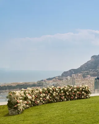 Installation de cérémonie de mariage en plein air avec des rangées de chaises dorées sur une pelouse, face à une composition florale incurvée de roses blanches et roses pâles, avec vue sur la mer et les montagnes.