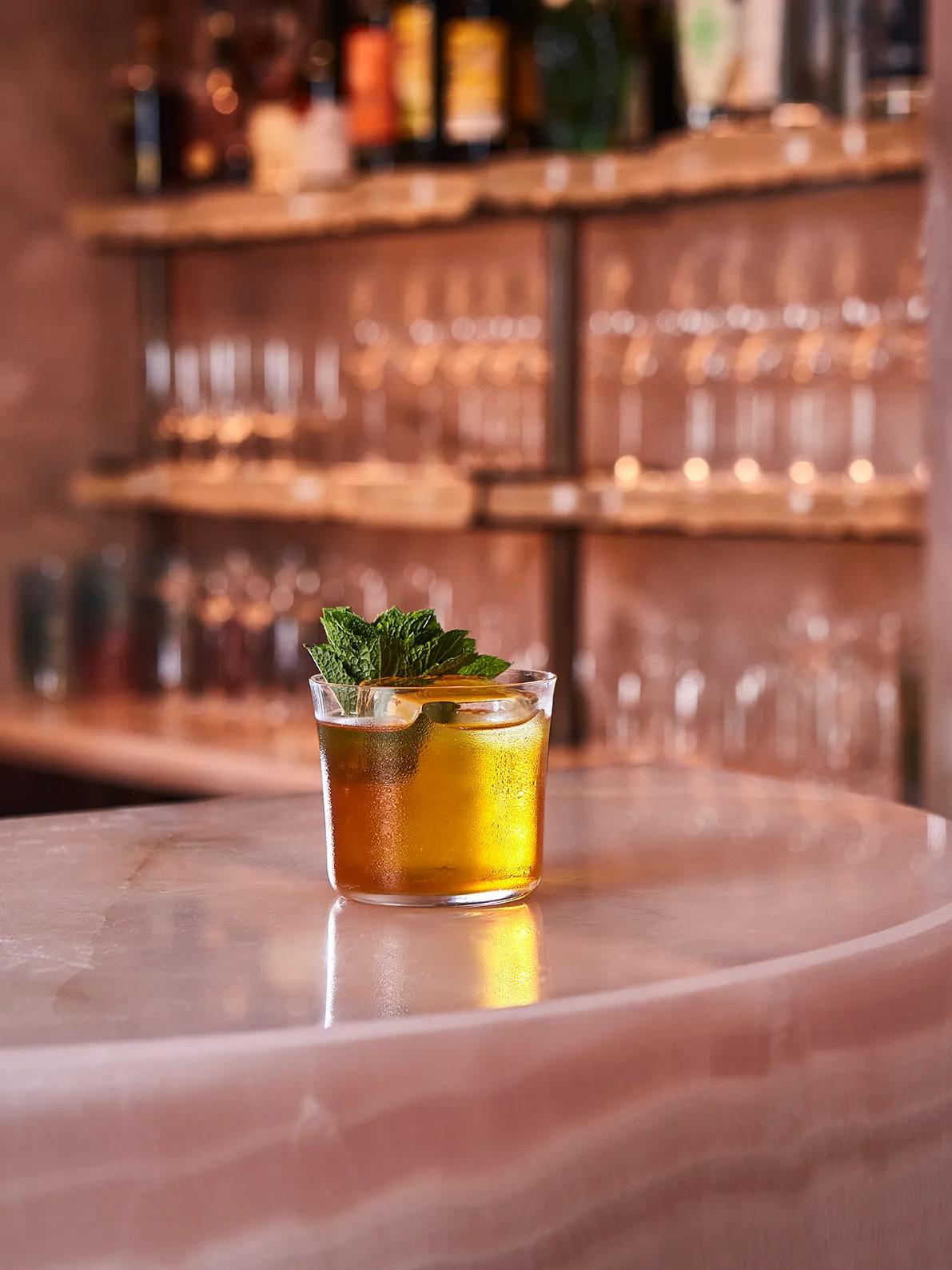 Amber cocktail garnished with mint and citrus slices, served on a marble bar counter with glassware behind.
