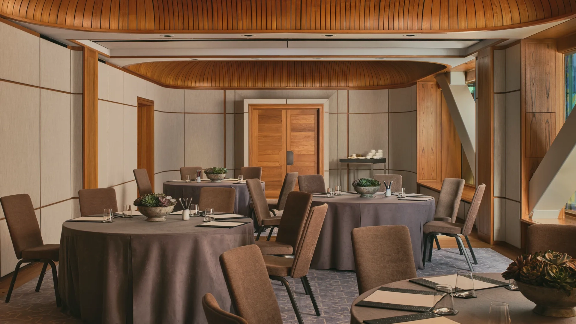 Boardroom setup with grey tablecloth, neutral chairs, succulents, and colourful abstract window panels under a curved wooden ceiling.