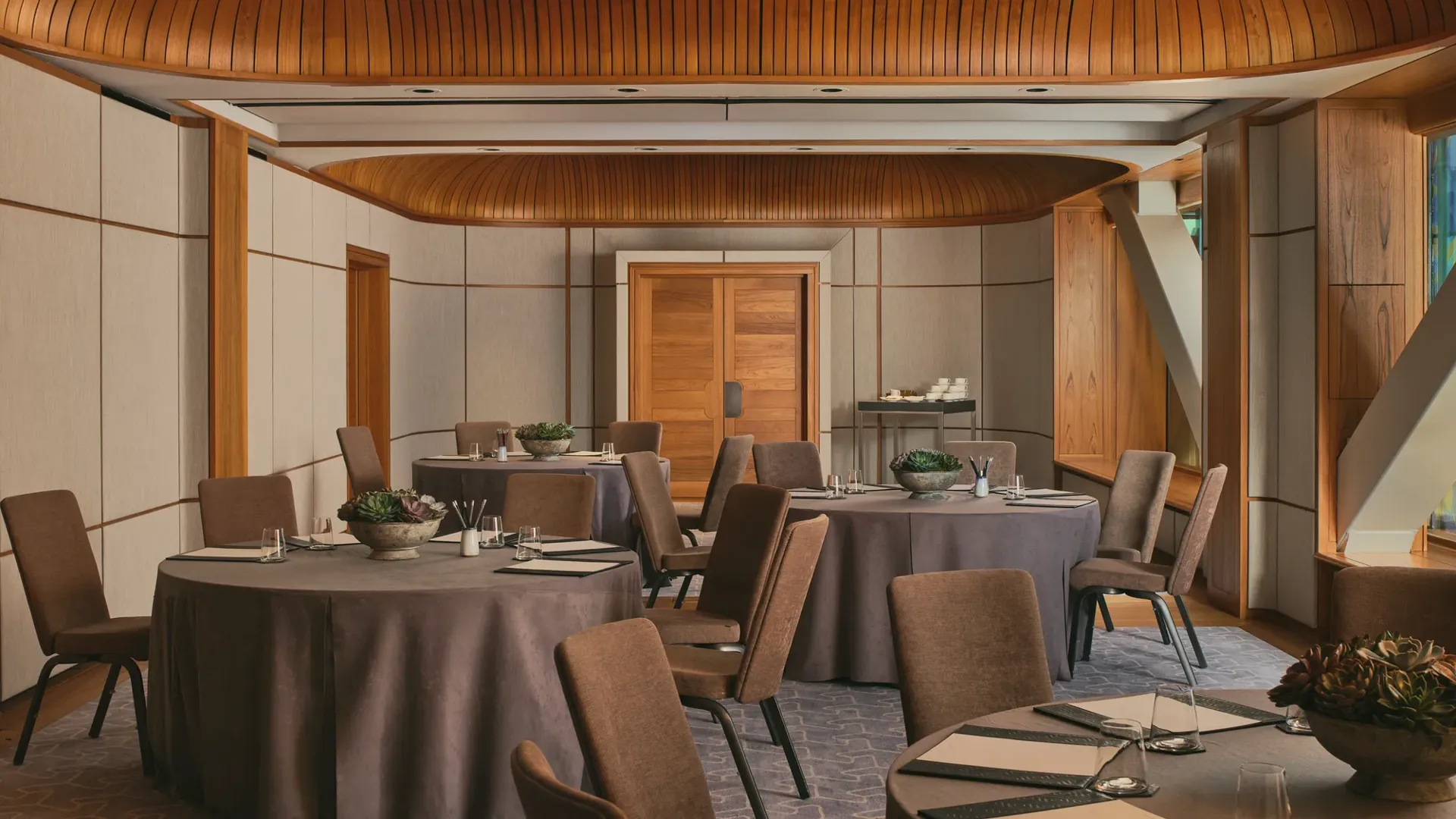 Boardroom setup with grey tablecloth, neutral chairs, succulents, and colourful abstract window panels under a curved wooden ceiling.