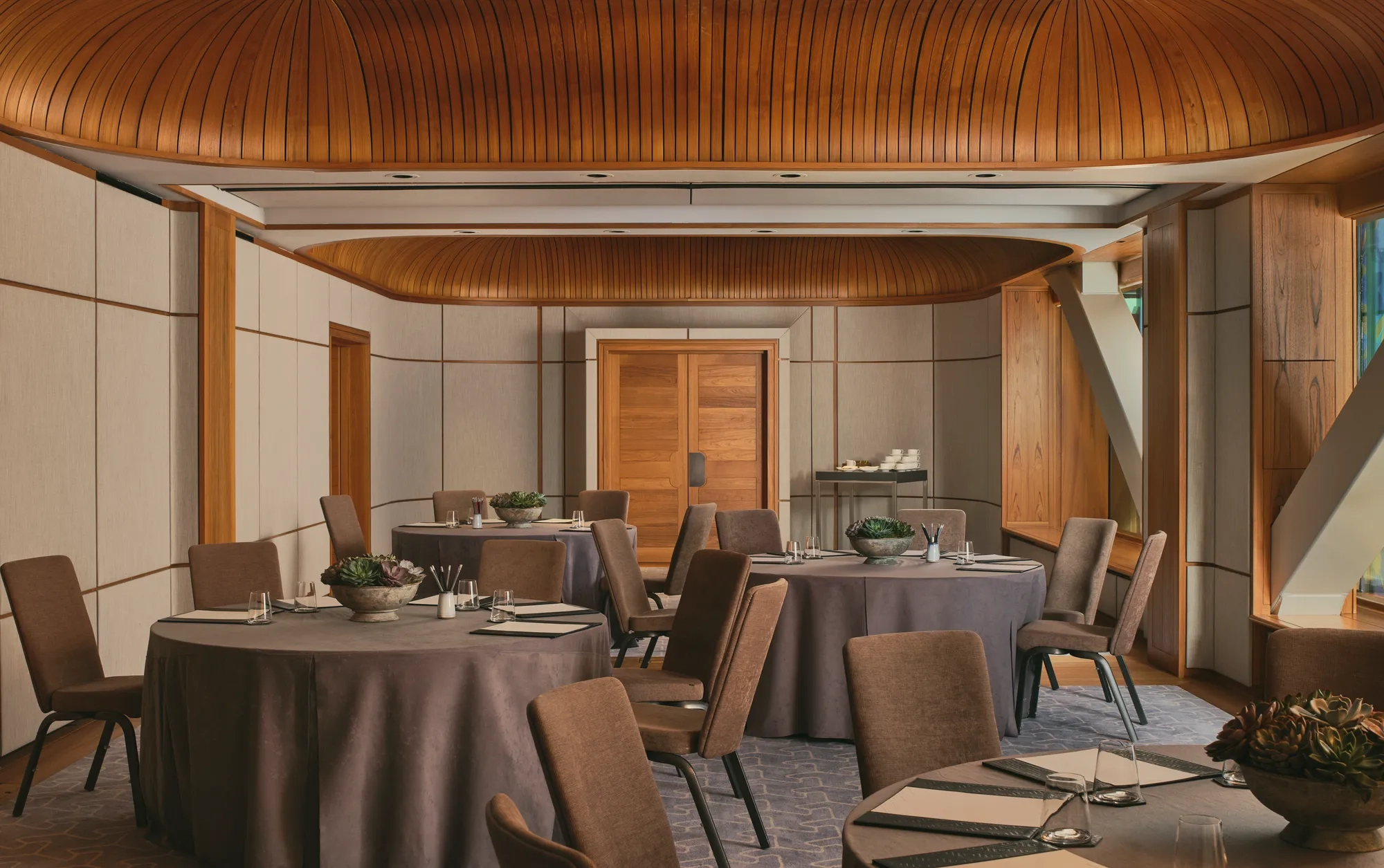 Boardroom setup with grey tablecloth, neutral chairs, succulents, and colourful abstract window panels under a curved wooden ceiling.