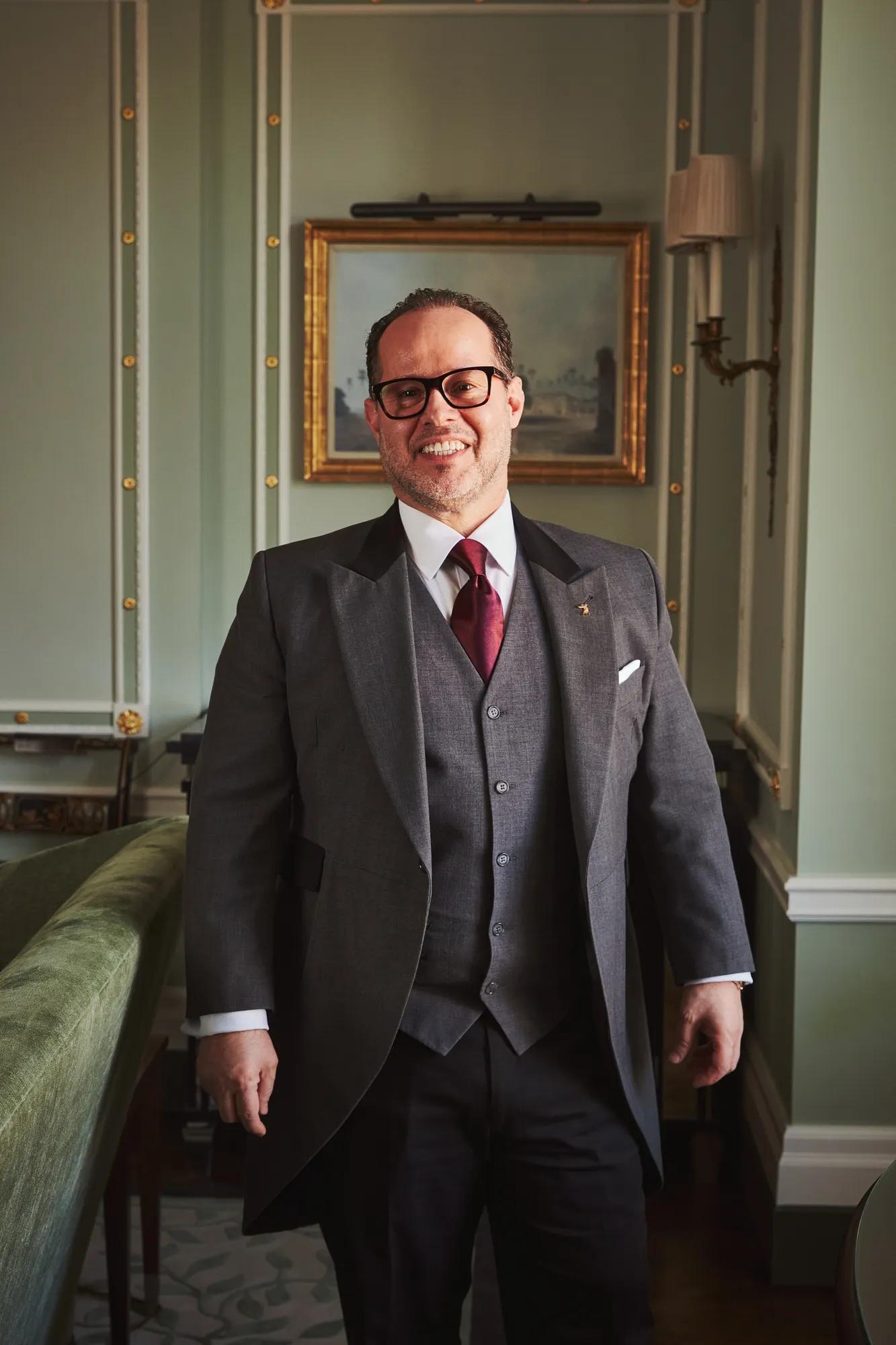 Smartly dressed hotel butler in a grey tailcoat and red tie stands smiling in an elegant, light green room.
