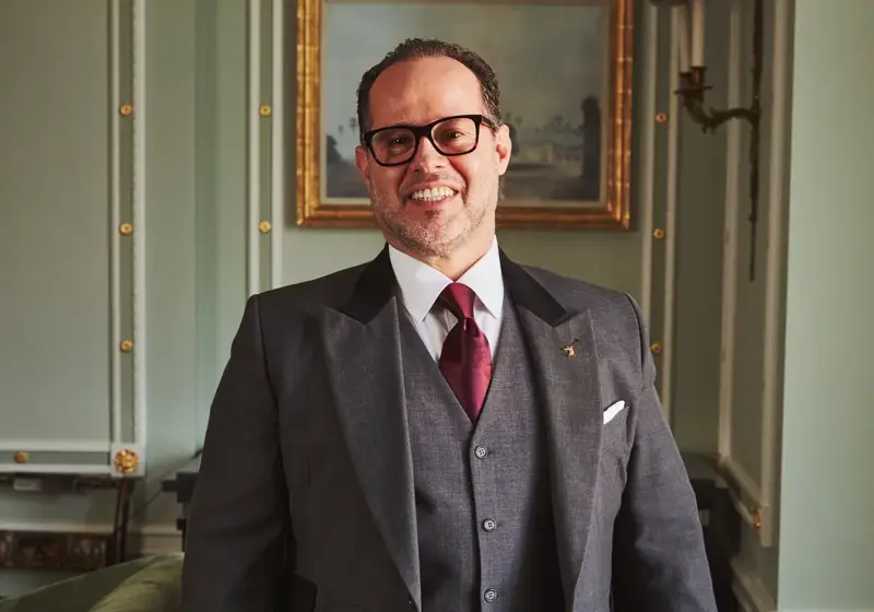 Smartly dressed hotel butler in a grey tailcoat and red tie stands smiling in an elegant, light green room.
