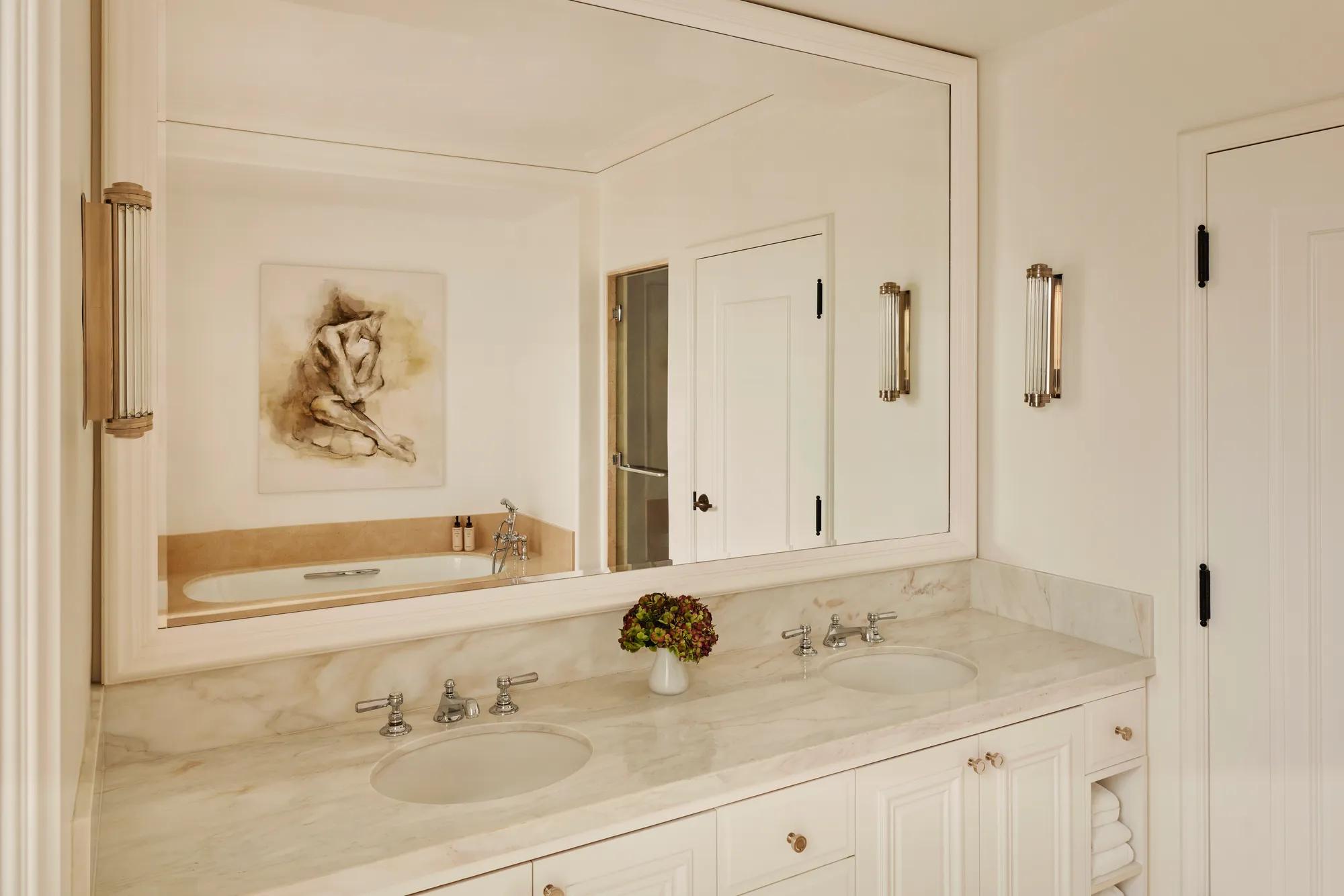 Marble bathroom with dual sinks, large framed mirror, and a bathtub reflected in the glass, accented with wall lights and a small vase of flowers.