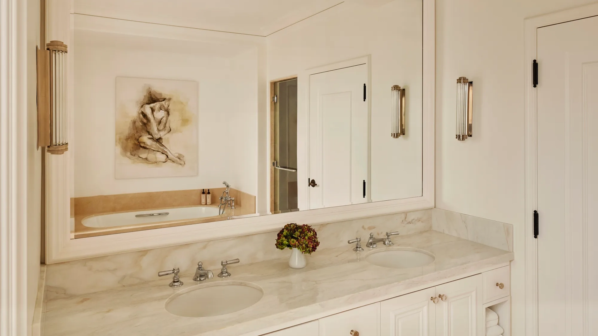 Marble bathroom with dual sinks, large framed mirror, and a bathtub reflected in the glass, accented with wall lights and a small vase of flowers.