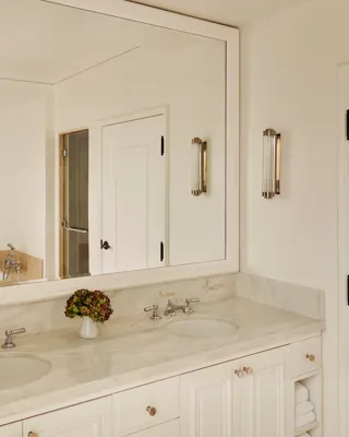 Marble bathroom with dual sinks, large framed mirror, and a bathtub reflected in the glass, accented with wall lights and a small vase of flowers.