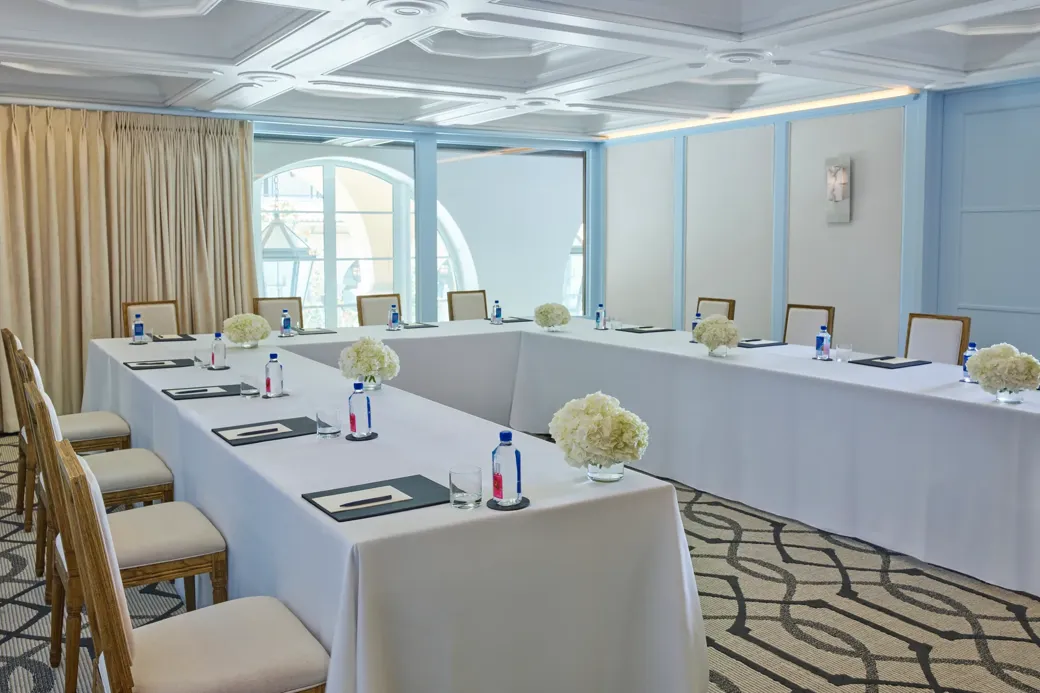 Beverly room in U-shape meeting setup with draped tables, water and notepads, blue paneling, and arched window.