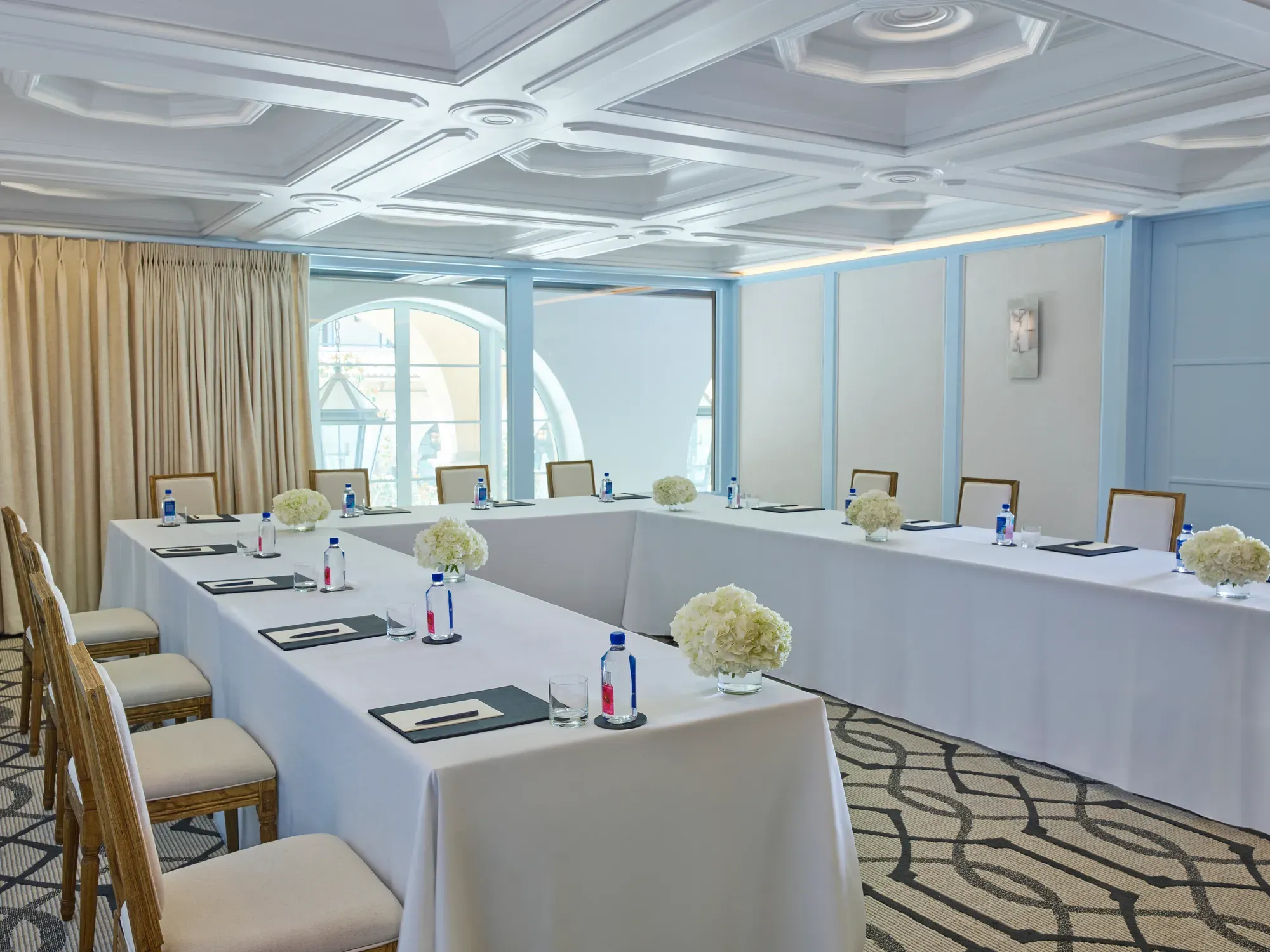 Beverly room in U-shape meeting setup with draped tables, water and notepads, blue paneling, and arched window.