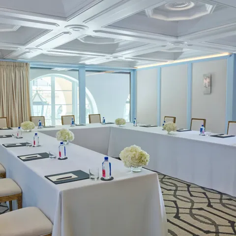 Beverly room in U-shape meeting setup with draped tables, water and notepads, blue paneling, and arched window.
