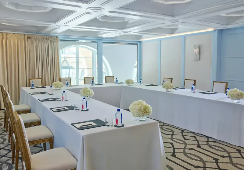 Beverly room in U-shape meeting setup with draped tables, water and notepads, blue paneling, and arched window.
