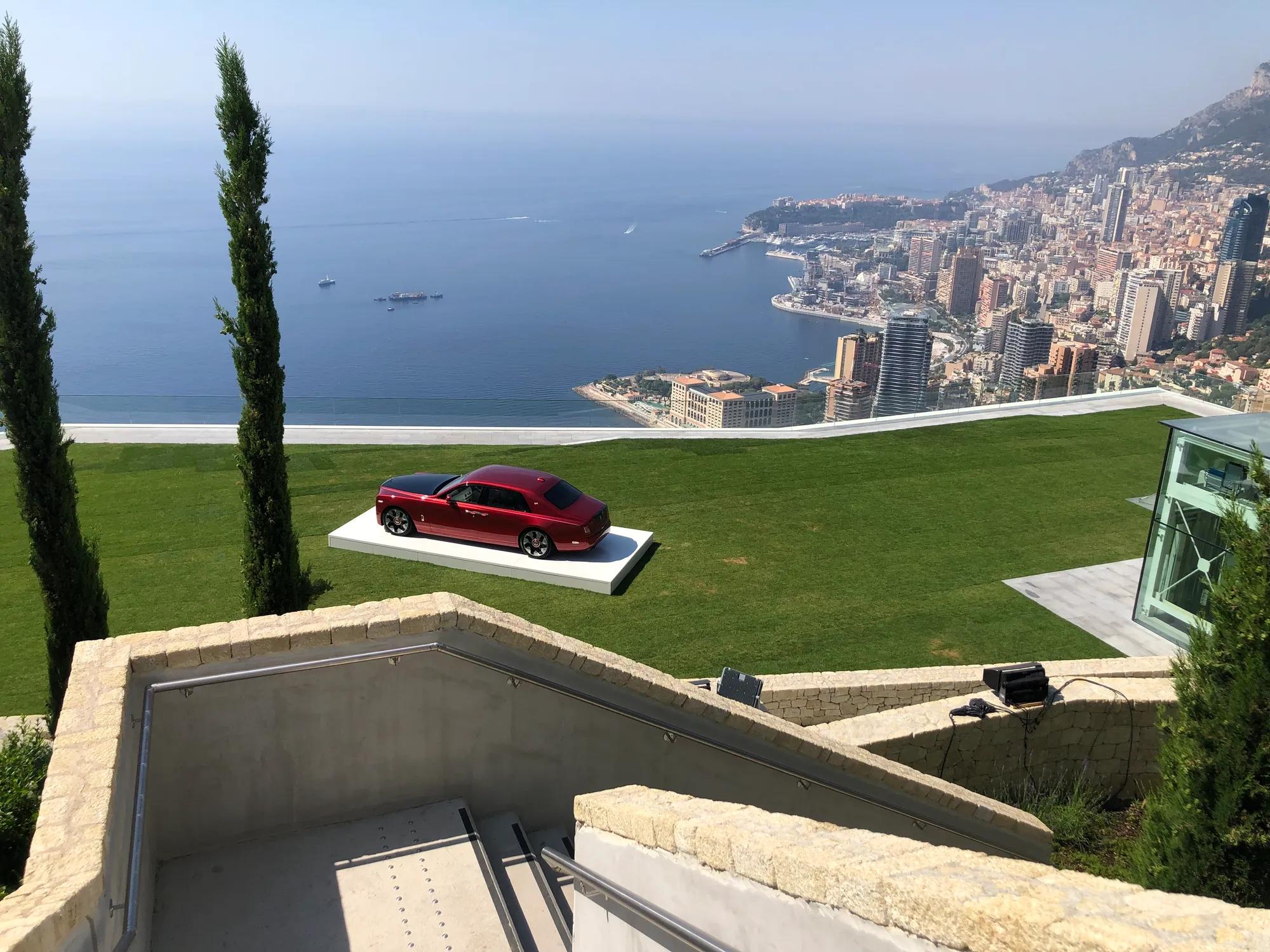 Red luxury car displayed on a lawn overlooking a coastal city and sea view.