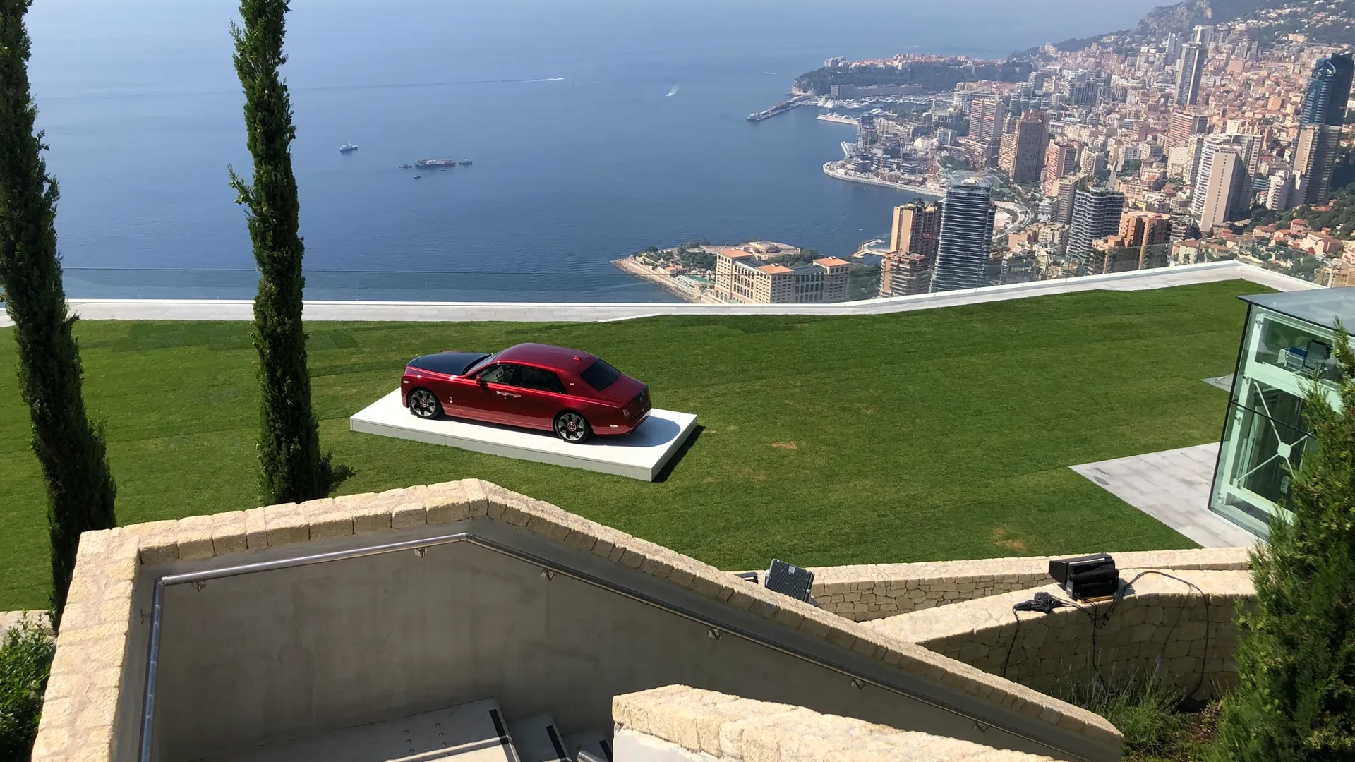 Voiture de luxe rouge exposée sur pelouse dominant une ville côtière et la mer.
