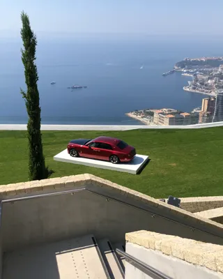 Red luxury car displayed on a lawn overlooking a coastal city and sea view.