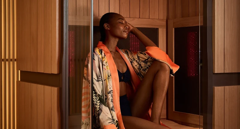 Person relaxing inside a wooden sauna cabin, wearing a robe and swimwear, seated on a bench with eyes closed.