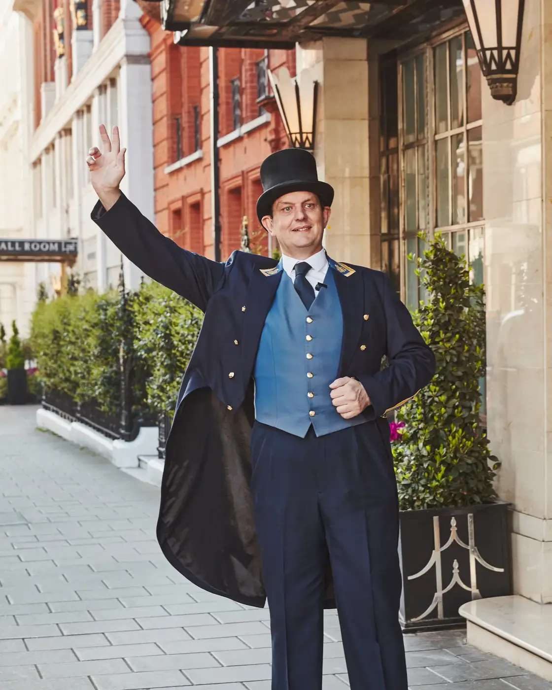 Claridge’s doorman in uniform and top hat waving outside the hotel entrance on Brook Street.