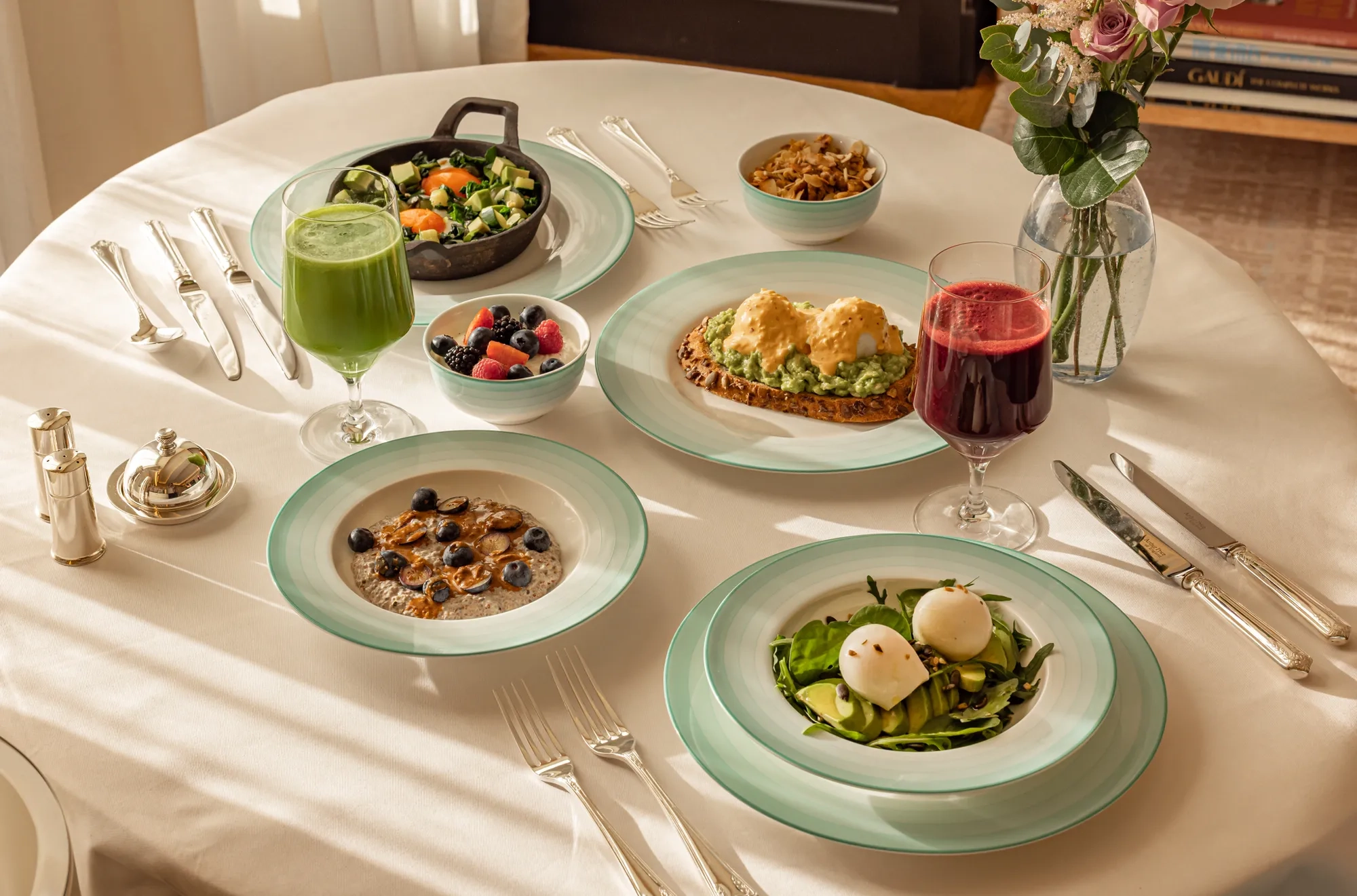 Healthy breakfast spread with eggs, avocado toast, fruit, pancakes, and fresh juices on a white tablecloth.