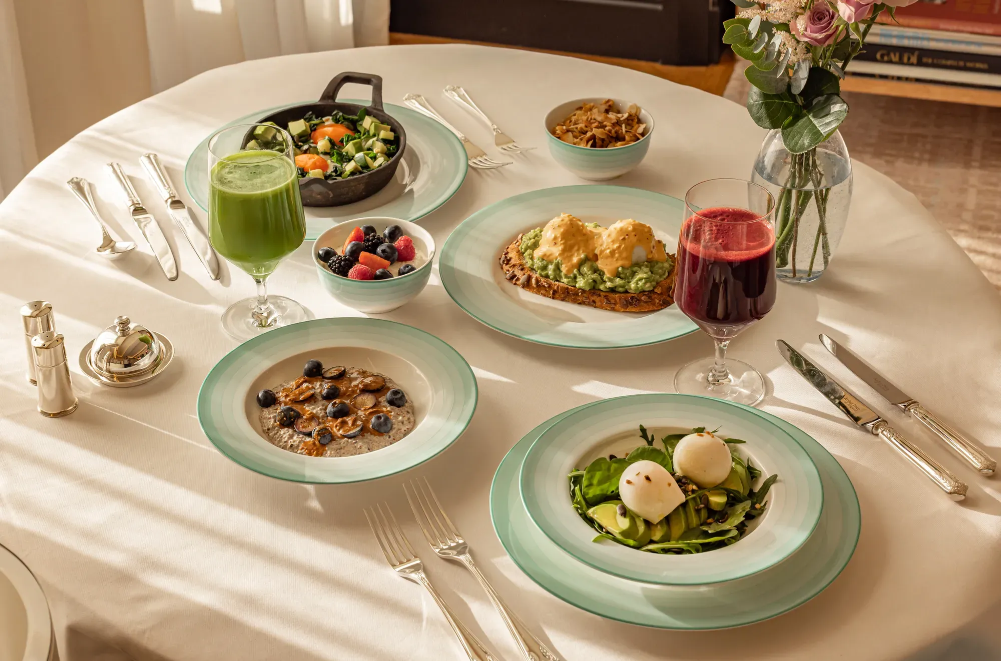Healthy breakfast spread with eggs, avocado toast, fruit, pancakes, and fresh juices on a white tablecloth.