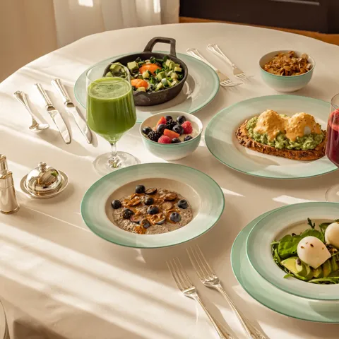Healthy breakfast spread with eggs, avocado toast, fruit, pancakes, and fresh juices on a white tablecloth.