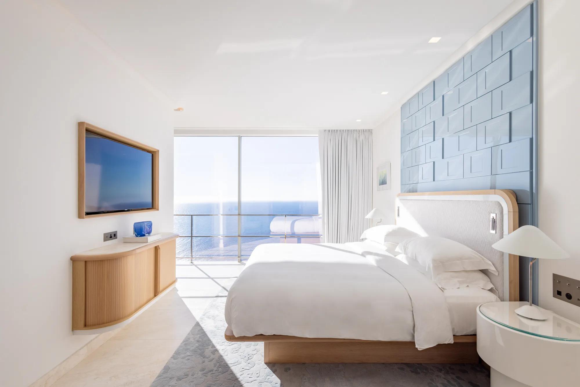 Chambre d’hôtel lumineuse avec grand lit, mur d’accent bleu et portes vitrées s’ouvrant sur une vue panoramique sur la mer, complétée par une télévision murale.