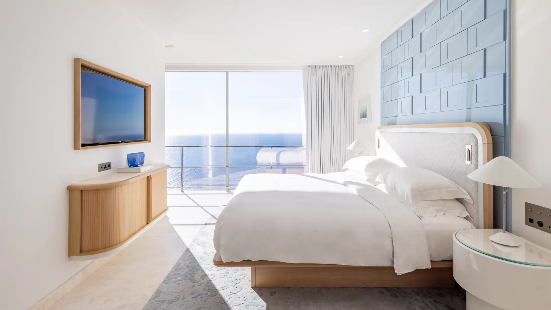 Chambre d’hôtel lumineuse avec grand lit, mur d’accent bleu et portes vitrées s’ouvrant sur une vue panoramique sur la mer, complétée par une télévision murale.