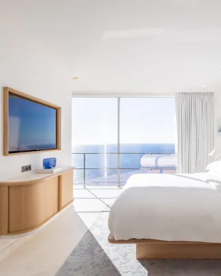 Chambre d’hôtel lumineuse avec grand lit, mur d’accent bleu et portes vitrées s’ouvrant sur une vue panoramique sur la mer, complétée par une télévision murale.