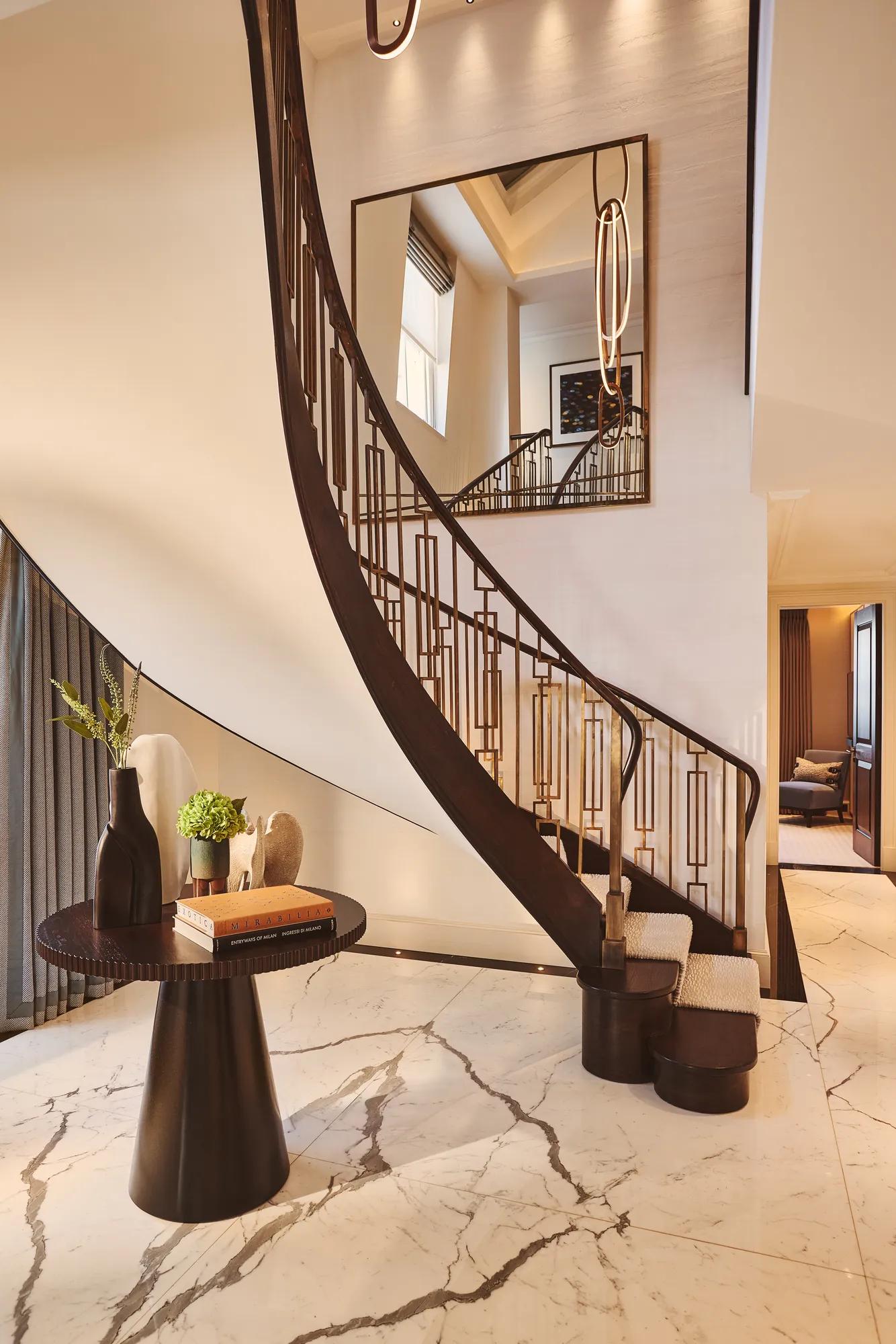 Curved staircase with dark wood railing in a marble-floored suite, beside a sculptural table and large wall mirror.