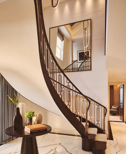 Curved staircase with dark wood railing in a marble-floored suite, beside a sculptural table and large wall mirror.