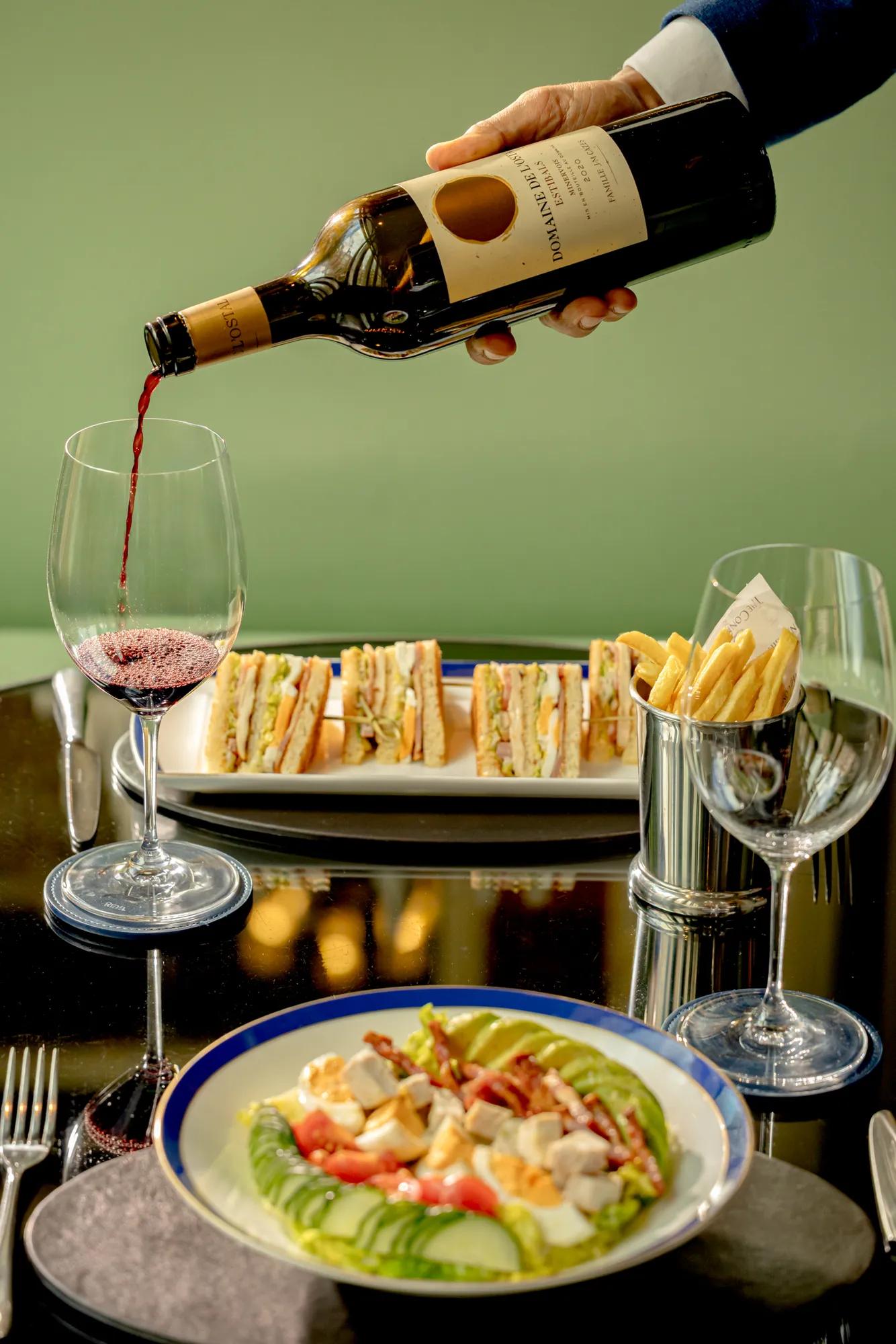 Red wine being poured into a glass beside a table set with a salad, sandwiches, and fries.