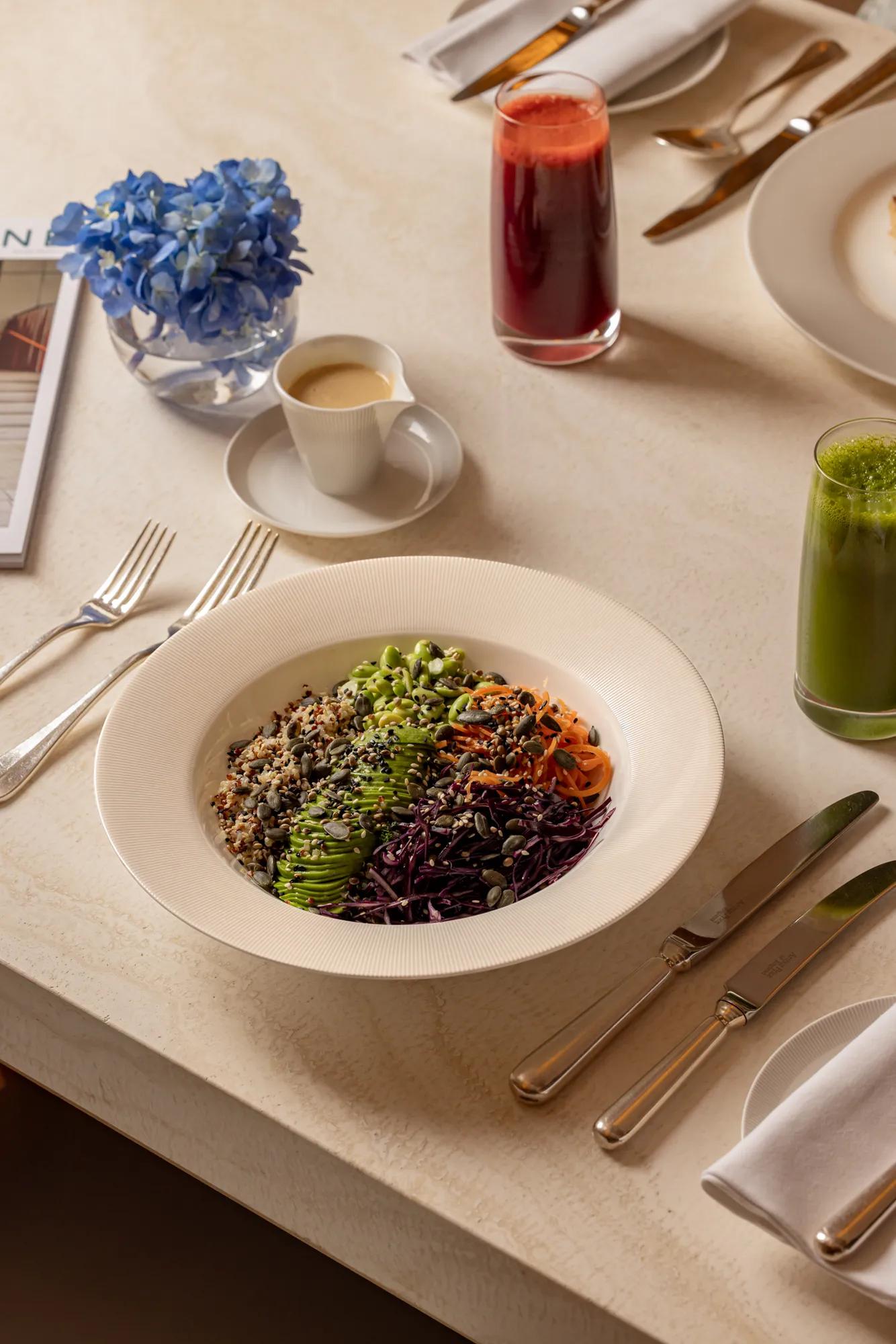 Colourful grain and vegetable bowl with juices and espresso on a marble table, beside blue flowers and neatly set cutlery.