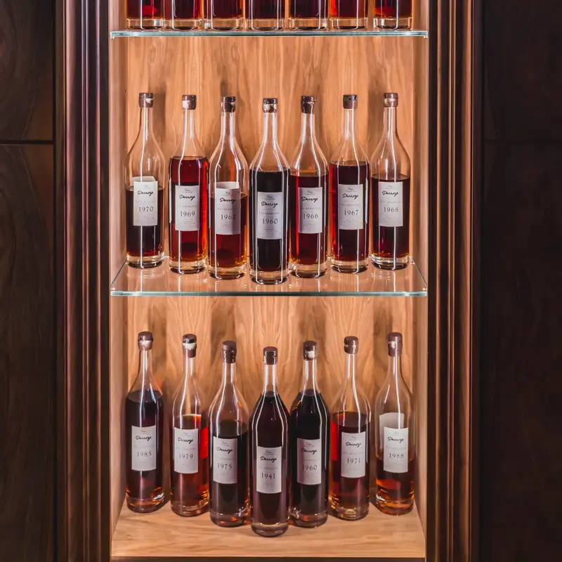 Illuminated wooden display cabinet with glass shelves holding rows of vintage cognac bottles with white labels and amber tones