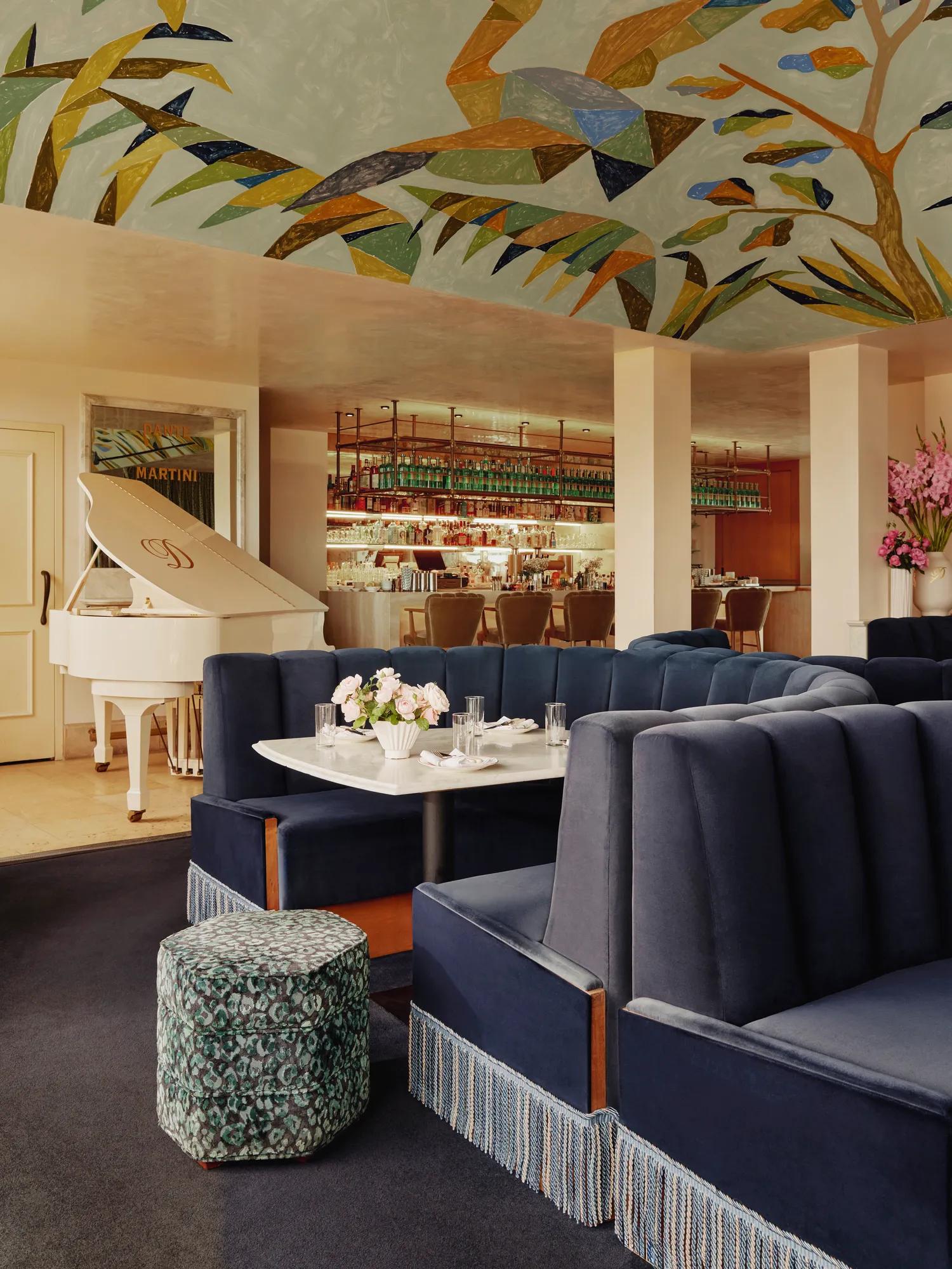 Dante Beverly Hills restaurant and rooftop with navy velvet banquettes and fringe, white grand piano, and a colorful ceiling mural near the bar.