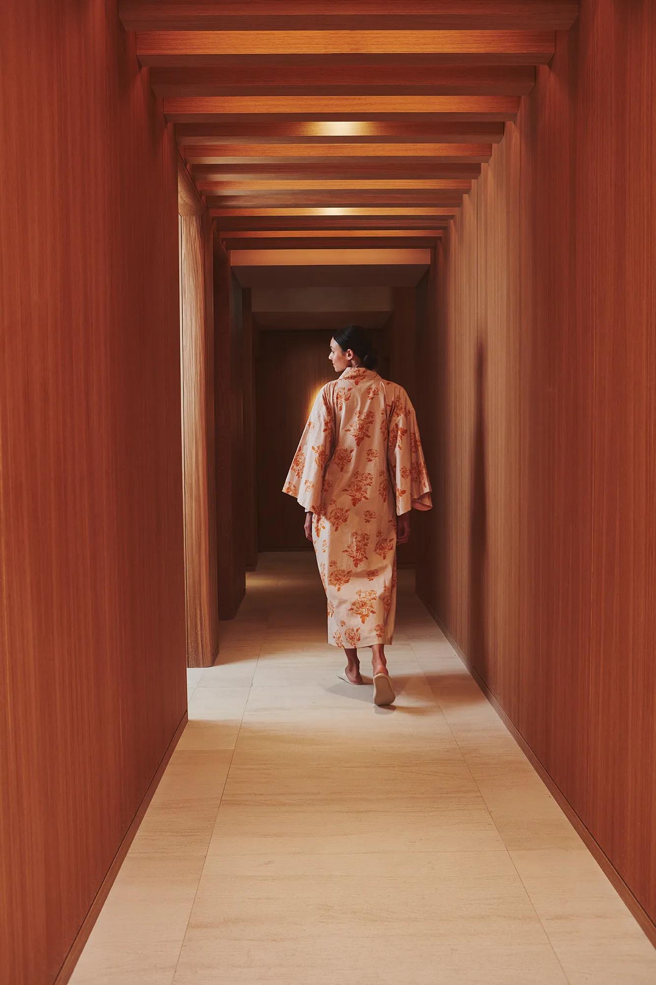 A woman wearing a patterned kimono walks down a softly lit wooden corridor with a geometric ceiling at The Maybourne Riviera.