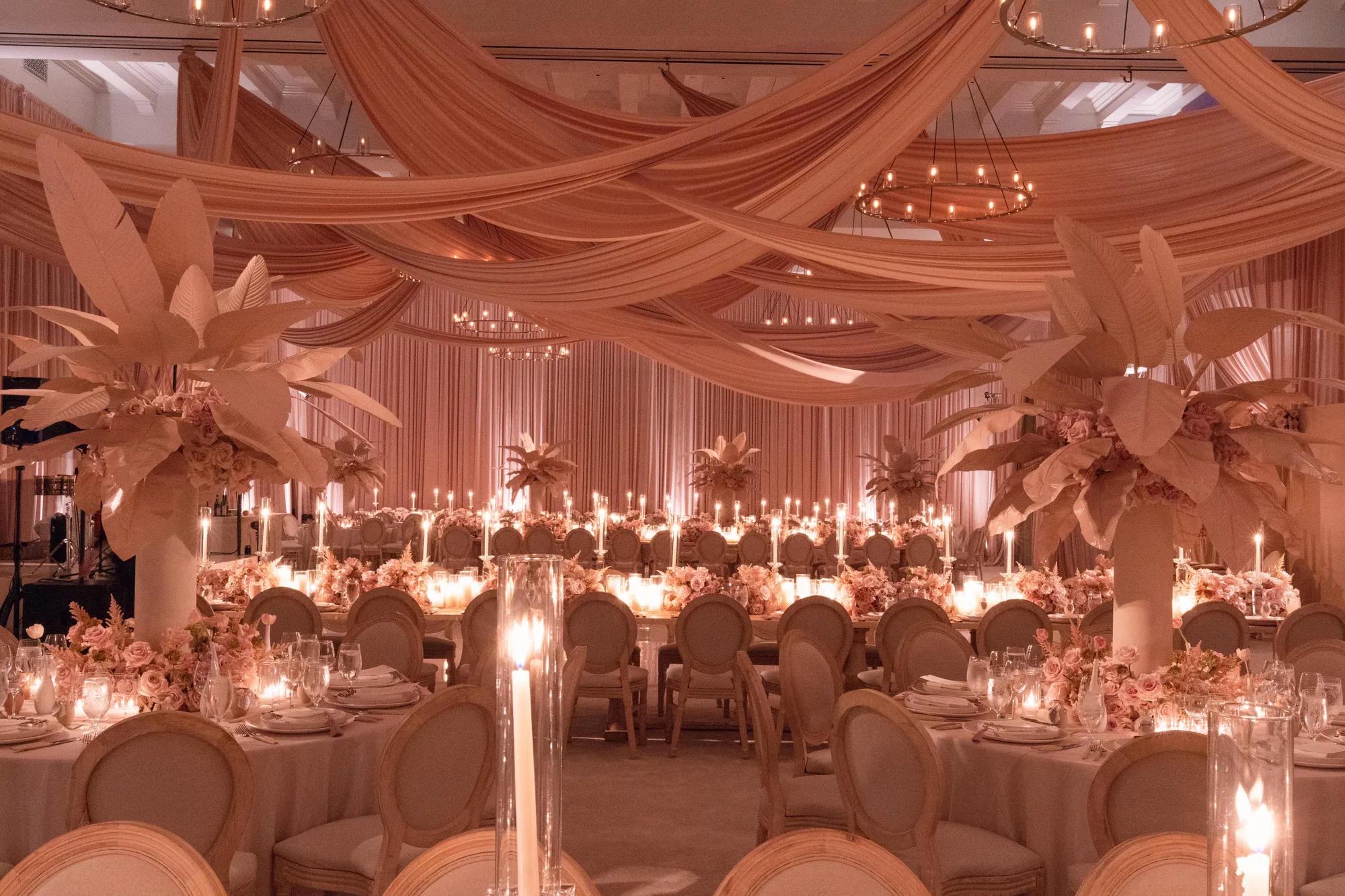 A softly lit wedding reception room decorated in blush tones, with draped peach fabric across the ceiling, tall floral centrepieces with pale leaves, round tables set with candles and fine glassware, and long banquet tables lined with flickering candlelight.
