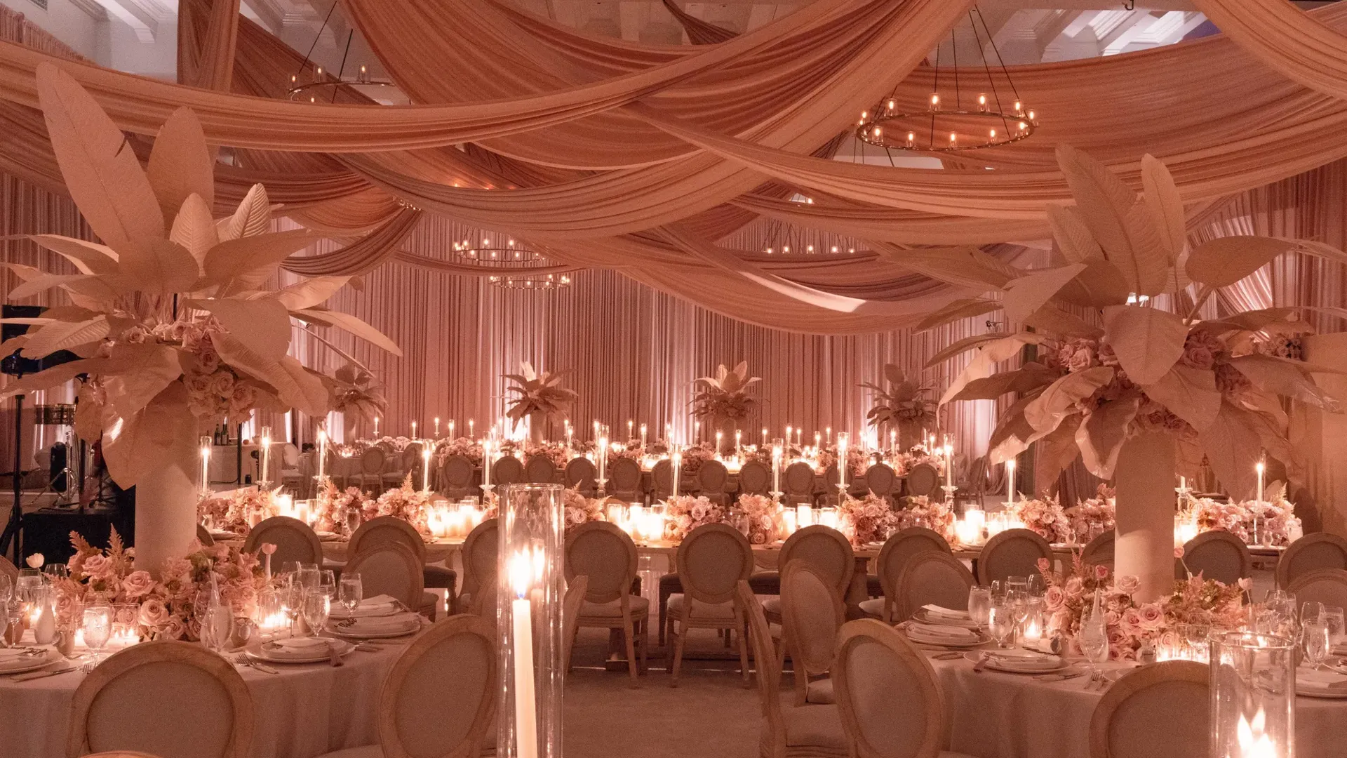 A softly lit wedding reception room decorated in blush tones, with draped peach fabric across the ceiling, tall floral centrepieces with pale leaves, round tables set with candles and fine glassware, and long banquet tables lined with flickering candlelight.