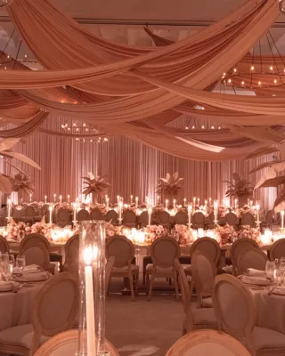 A softly lit wedding reception room decorated in blush tones, with draped peach fabric across the ceiling, tall floral centrepieces with pale leaves, round tables set with candles and fine glassware, and long banquet tables lined with flickering candlelight.