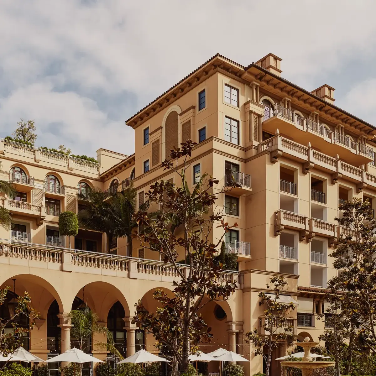 Arched terrace of The Maybourne Beverly Hills facing Beverly Canon Gardens, with palms and white umbrellas below.