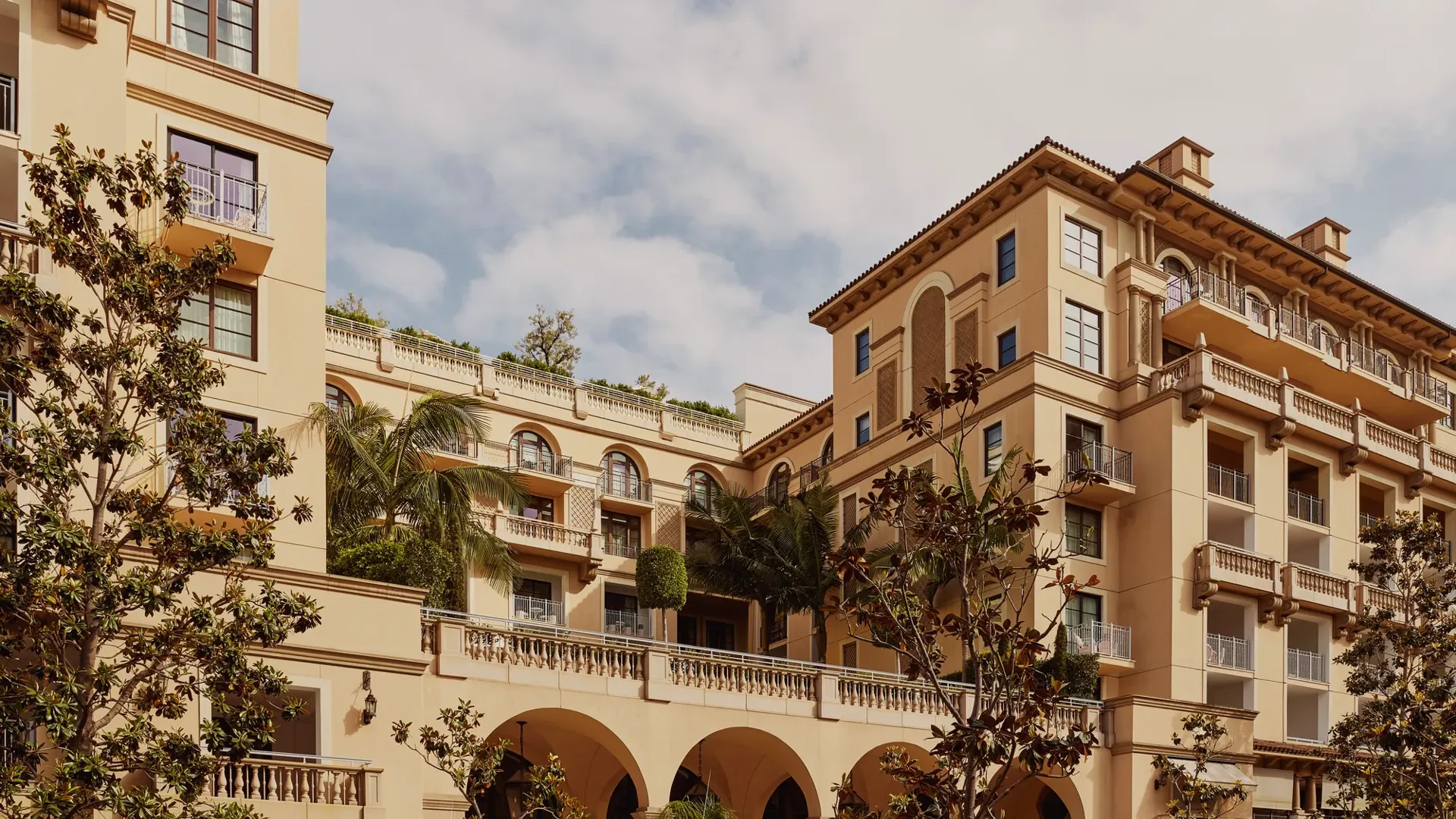 Arched terrace of The Maybourne Beverly Hills facing Beverly Canon Gardens, with palms and white umbrellas below.