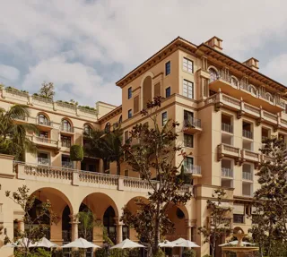Arched terrace of The Maybourne Beverly Hills facing Beverly Canon Gardens, with palms and white umbrellas below.